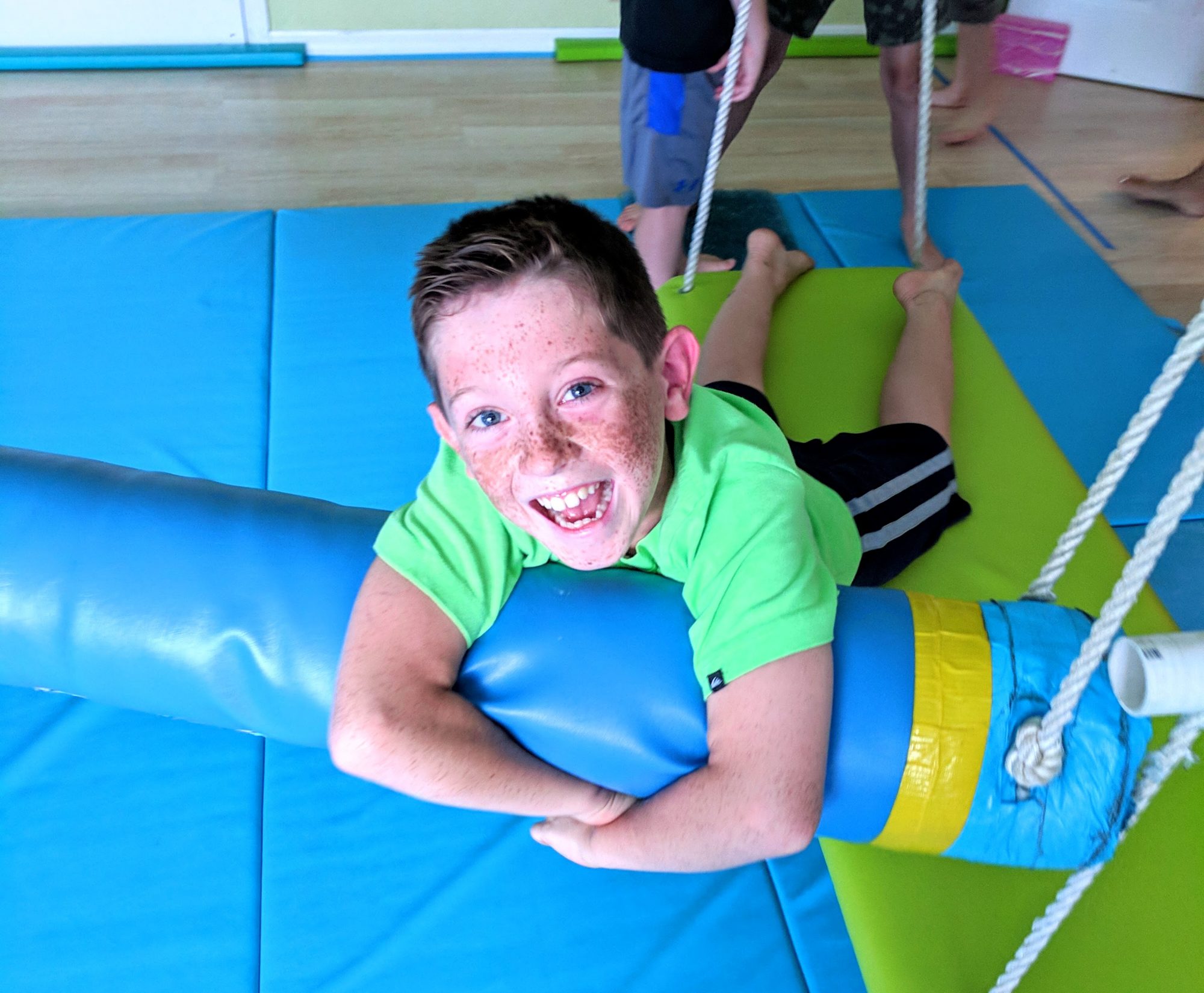 A young boy with freckles smiles joyfully while playfully hanging onto a large foam cylinder in a colorful therapy room, surrounded by soft mats.