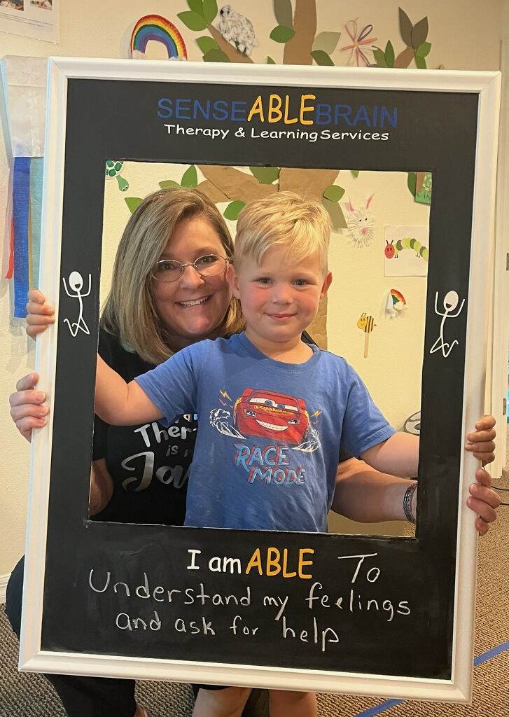 A smiling woman and a young boy pose together in a framed sign that reads 'I am ABLE To understand my feelings and ask for help' at Sense Able Brain Therapy & Learning Services, surrounded by colorful educational decorations.